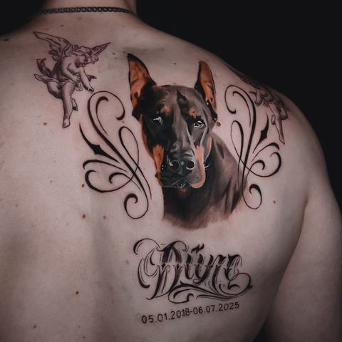 A memorial back tattoo featuring a realistic Doberman portrait, ornamental lines, cherubs, a name and dates, creating a deeply personal composition.