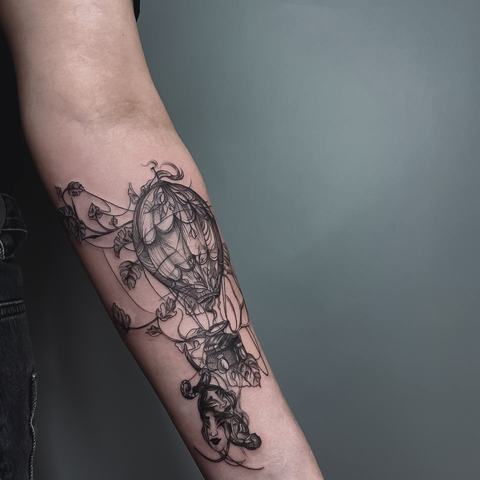 A detailed black-and-grey forearm tattoo with an ornate balloon-like form, floral elements and fine shading that creates a layered, illustrative composition.