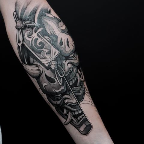 A striking mask tattoo on the forearm with flowing ornamental shapes, deep black areas and controlled shading that gives the piece a powerful sculptural effect.
