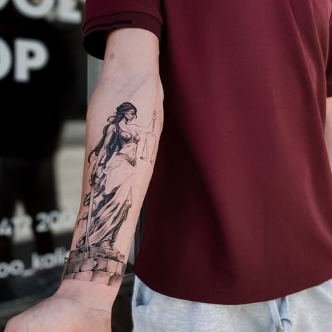 An elegant Lady Justice tattoo on the forearm, featuring scales and flowing fabric. Fine lines and soft shading give the figure movement and balance.