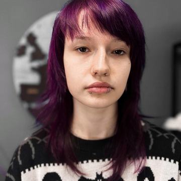 Portrét osoby s fialovými vlasmi a dvoma piercingmi v spodnej pere (krúžok a labret), v čierno-bielom svetri s motívom netopierov; ilustračné foto k profesionálnemu piercingu pier v štúdiu VEAN TATTOO v Prešove.