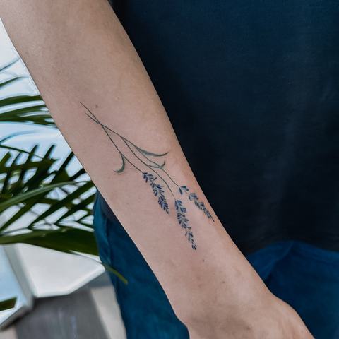 A minimalist lavender tattoo on the forearm, combining thin green stems with small blue-purple blossoms for a clean and delicate botanical composition.