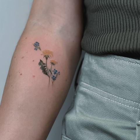A tiny wildflower tattoo with soft yellow and blue blossoms. Delicate stems and subtle colour make this small design feel natural, fresh and understated.