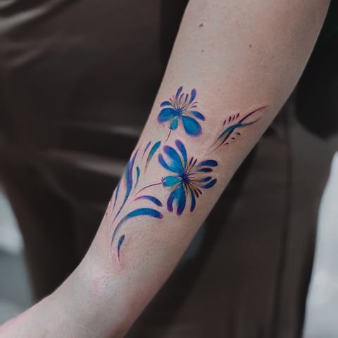 A bright blue floral tattoo with loose brush-like strokes and delicate details. The painterly style creates a fresh, artistic and lightweight composition.