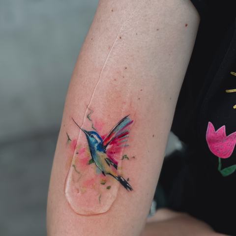 A colourful hummingbird tattoo in a soft watercolour style. Blue, pink and green tones blend with loose brush-like details for an airy, artistic effect.